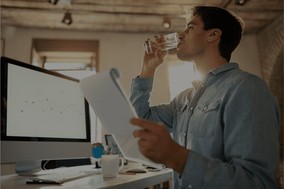 Ein junger Mann im Büro eines Unternehmens trinkt gekühltes Wasser aus einem Glas vor dem Computer und hält ein Papierblock in der linken Hand