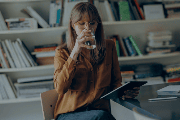 Eine Frau in einer Bibliothek trinkt während dem lesen ein Glas Wasser