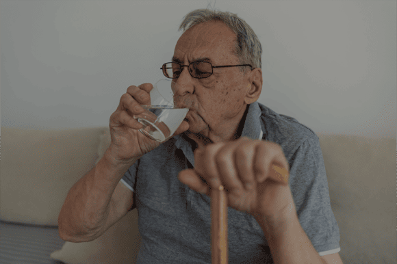 Ein alter Mann in einem Altersheim Pflegeheim trinkt Wasser aus einem Glas und hält sich am Gehstock